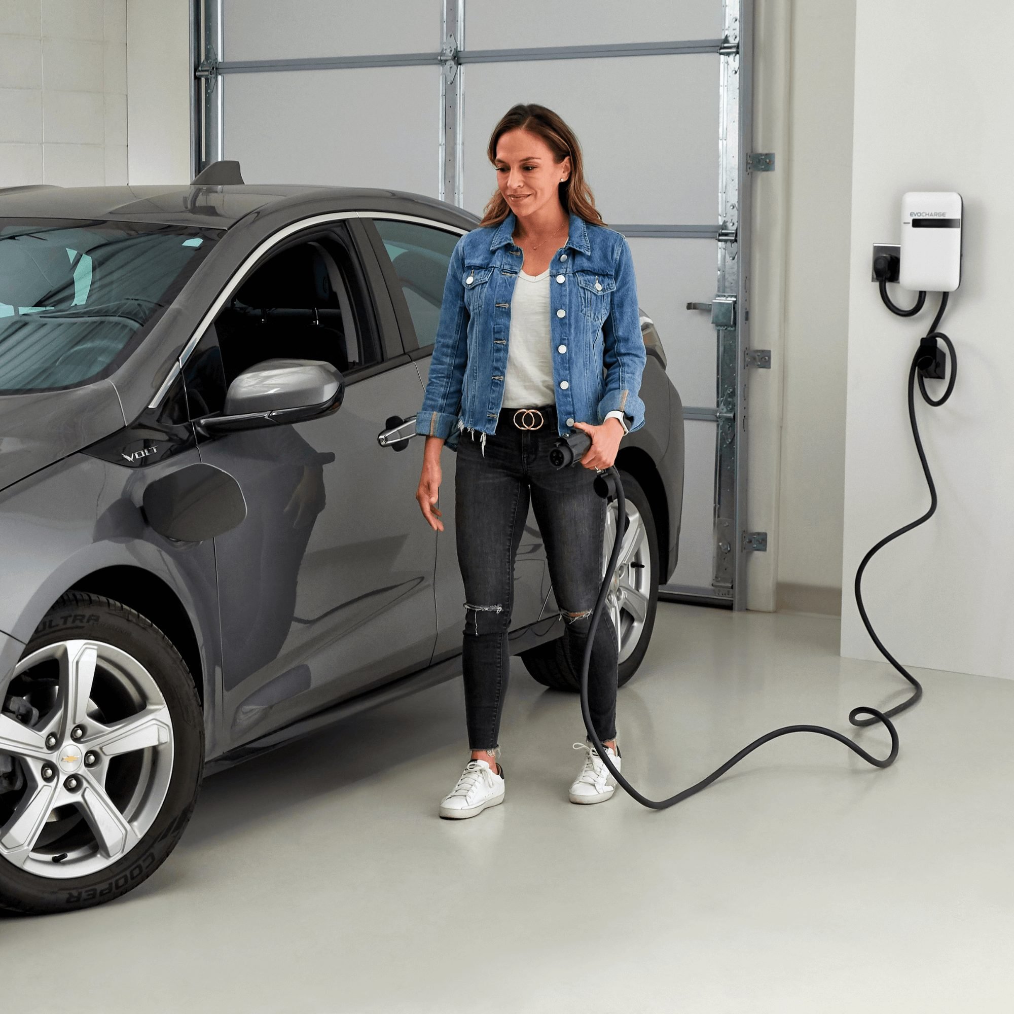 A woman in a jean jacket moving to plug in her electric vehicle in a garage