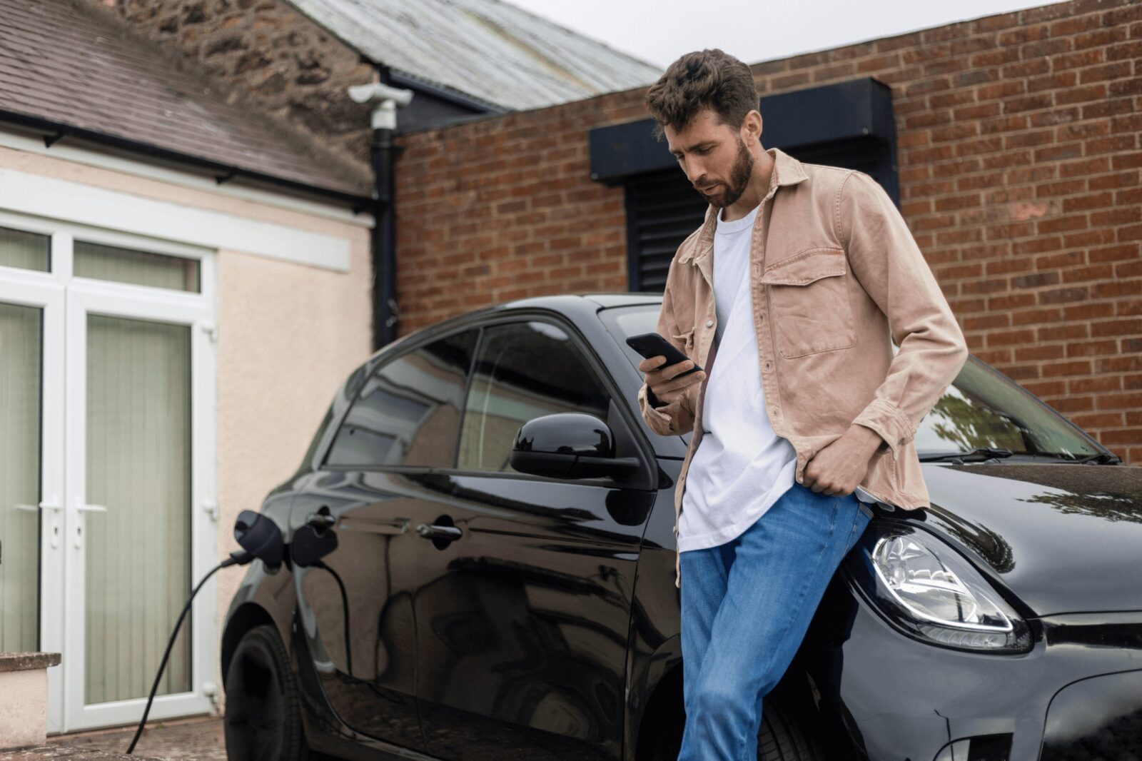 A man leans against his car, looking at his phone as his electric vehicle charges outside.